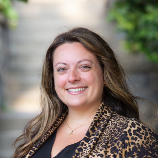 A woman with long brown hair smiles outdoors, wearing a leopard print jacket and a gold necklace. There's greenery and stone steps blurred in the background.